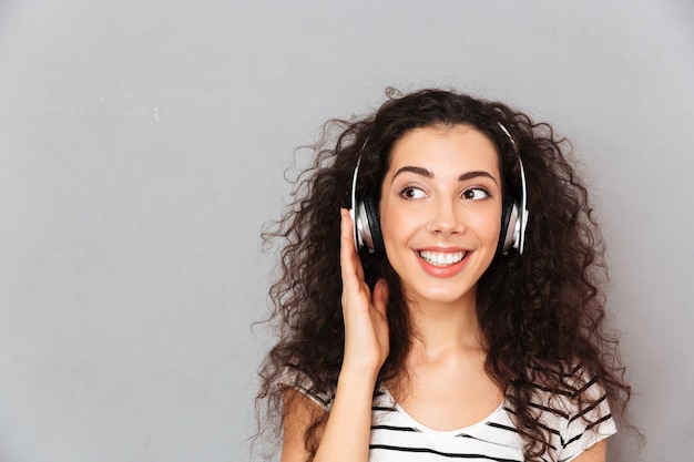 Großes Bild der gelockten kaukasischen Frau in gestreiftem T-Shirt in den Kopfhörern Musik über modernes Gerät beim Stillstehen genießend