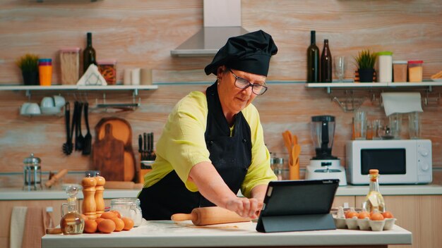 Großmutter hört Videotipps für die Zubereitung von hausgemachten Kuchen. Pensionierte Dame, die einen kulinarischen Podcast auf dem Tablet verfolgt und ein Koch-Tutorial in den sozialen Medien mit einem hölzernen Nudelholz lernt, das den Doug bildet