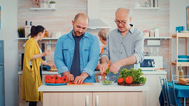 Großvater und Sohn im Esszimmer bereiten einen frischen Salat zu. Ein Mann mittleren Alters und ein älterer Senior haben Spaß daran, gemeinsam das Abendessen in einer modernen Küche zu kochen, während Frauen im Hintergrund sprechen