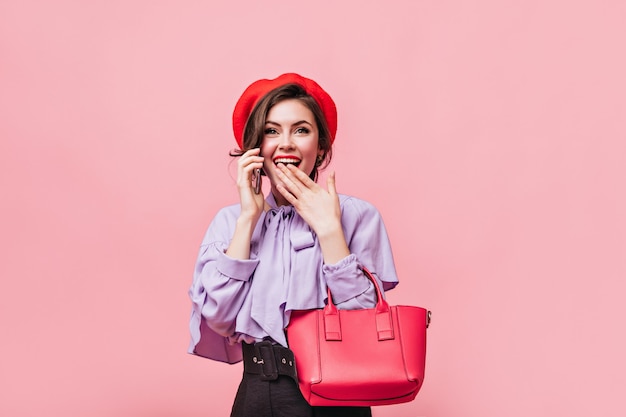 Grünäugige Frau hält sich den Mund zu und telefoniert. Dame in der roten Baskenmütze hält Tasche auf rosa Hintergrund.