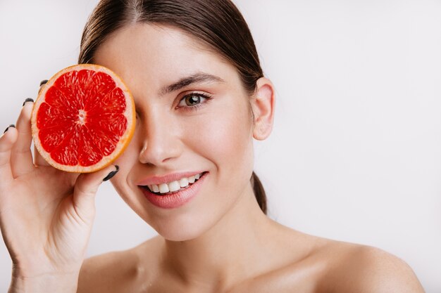 Grünäugige Frau mit Lächeln auf weißer Wand. Schnappschuss eines Modells ohne Make-up, das ihr Gesicht mit saftiger Grapefruit bedeckt.