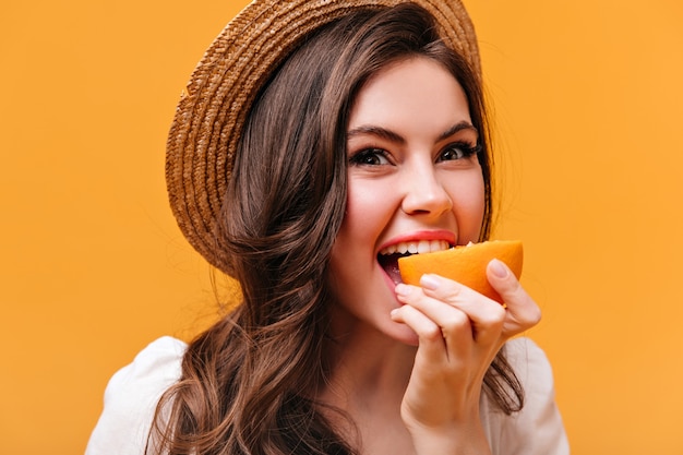 Grünäugiges brünettes Mädchen im Strohhut beißt saftige Orange auf lokalisiertem Hintergrund.