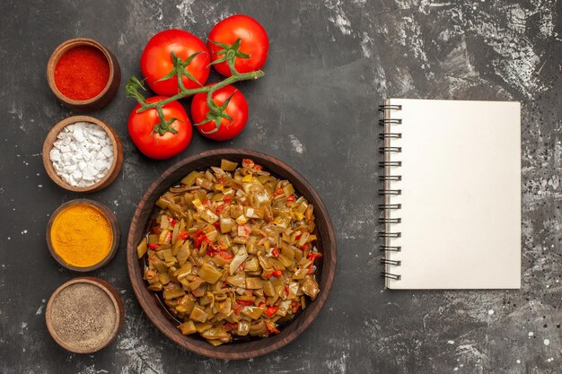 grüne Bohnen und Gewürze grüne Bohnen in der Schüssel neben dem weißen Notizbuch und die Schüsseln mit Gewürzen und Tomaten mit Stielen auf dem dunklen Tisch