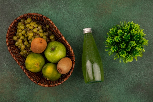 Grüne Mandarinen der Draufsicht mit Apfel-Kiwi und Trauben in einem Korb mit einer Flasche Saft auf einer grünen Wand