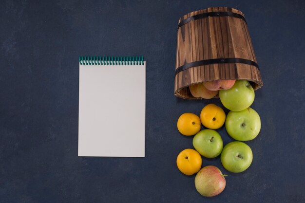 Grüne Äpfel und Orangen aus einem Holzeimer auf Schwarz mit einem Notizbuch beiseite
