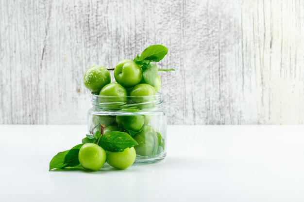 Grüne Pflaumen in einem Miniglas mit Seitenansicht der Blätter auf weißer und schmuddeliger Wand