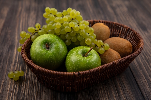 Grüne Trauben der Seitenansicht mit Äpfeln und Kiwi in einem Korb auf einem hölzernen Hintergrund