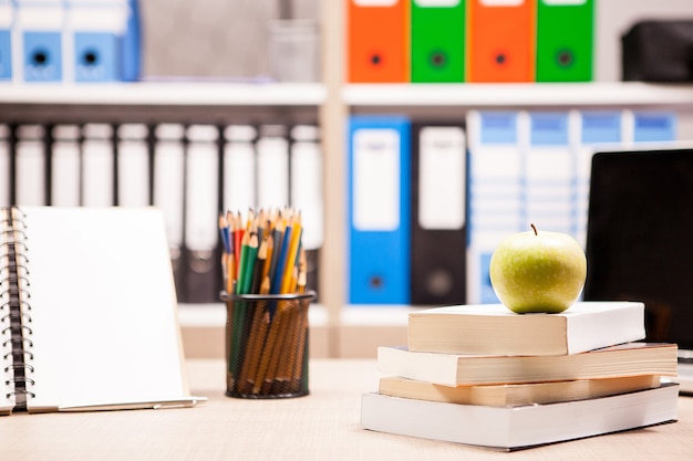 Grüner Apfel auf Bücherstapel neben einem Notizbuch und Bleistiften auf dem Tisch mit einem verschwommenen weißen Brett im Rücken. Schulkonzept