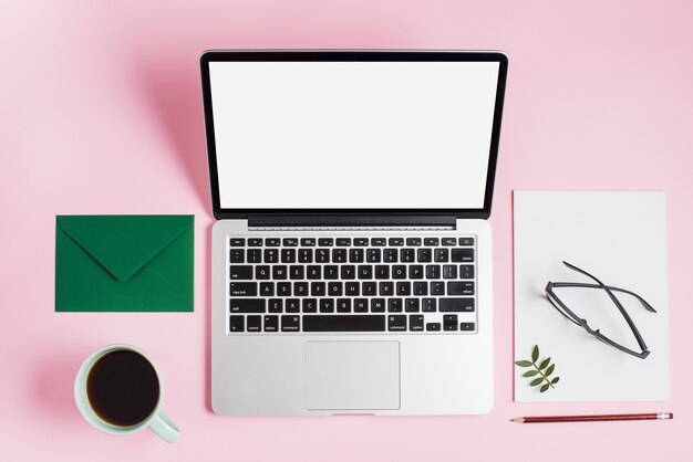 Grüner Umschlag; Teetasse; Laptop; Brillen auf Papier und Bleistift auf rosa Hintergrund