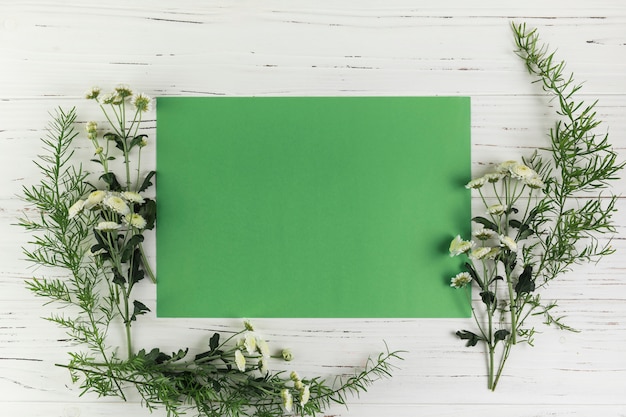 Grünes leeres Papier mit Chrysanthemenblumen und -blättern auf weißem hölzernem Schreibtisch