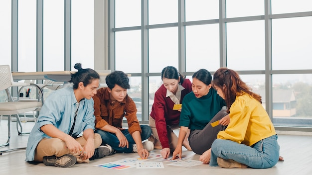 Gruppe asiatischer junger kreativer Leute in der Freizeitkleidung, die Geschäfts-Brainstorming-Besprechungsideen diskutiert, die Designplan des mobilen Anwendungssoftwareentwurfs auf Boden im Büro angelegt werden. Teamwork-Konzept für Mitarbeiter.