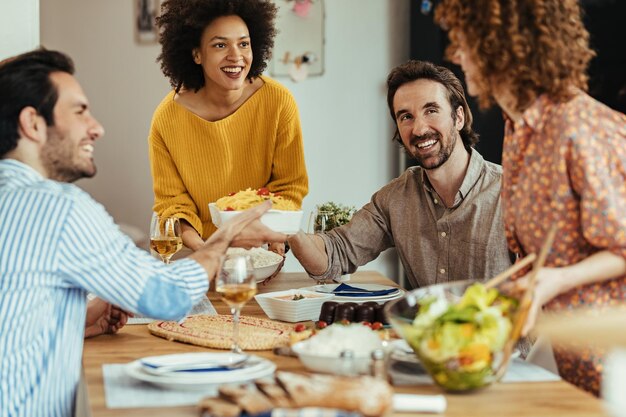 Gruppe glücklicher Freunde, die Essen am Tisch bringen und kommunizieren, während sie zu Hause zu Mittag essen