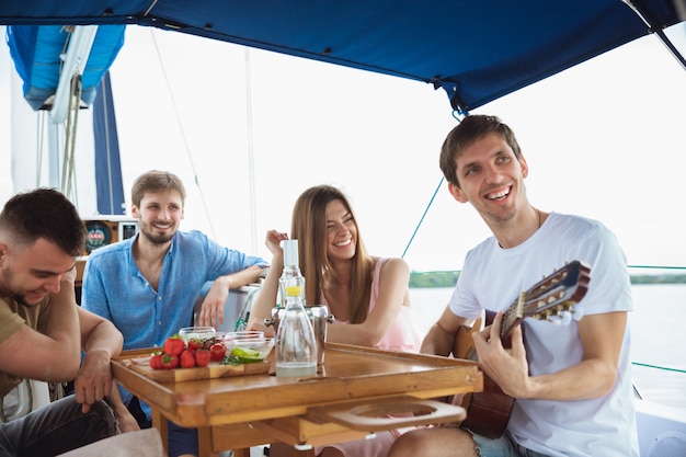Gruppe glücklicher Freunde, die Wodka-Cocktails trinken und Gitarre in einem Boot spielen