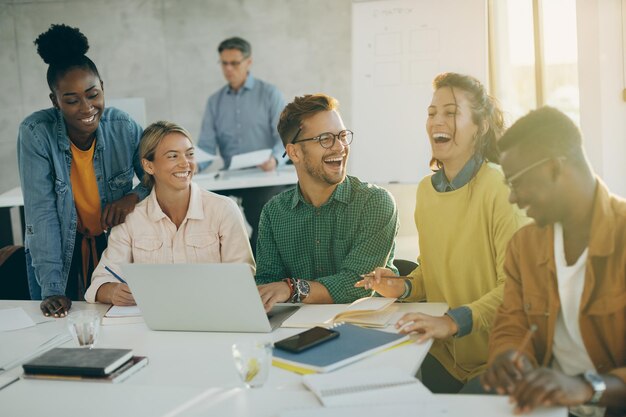 Gruppe glücklicher Studenten, die Spaß haben und lachen, während sie Laptops benutzen und im Klassenzimmer lernen. Ihr Lehrer ist im Hintergrund.