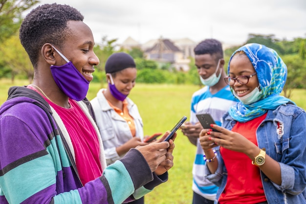 Gruppe junger afrikanischer Freunde mit Gesichtsmasken, die ihre Telefone in einem Park benutzen