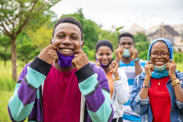 Gruppe junger fröhlicher afrikanischer Freunde, die Gesichtsmasken und soziale Distanzierung in einem Park tragen