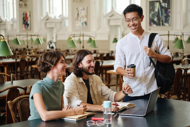 Gruppe junger, fröhlicher Studenten, die sich freudig unterhalten, während sie gemeinsam in der Bibliothek der Universität studieren