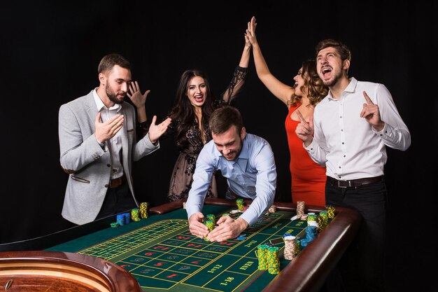 Gruppe junger Leute hinter Roulette-Tisch auf schwarzem Hintergrund. Gewinnender Spieler. Helle Emotionen