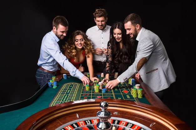 Gruppe junger Leute hinter Roulette-Tisch auf schwarzem Hintergrund. Junge Leute wetten im Spiel