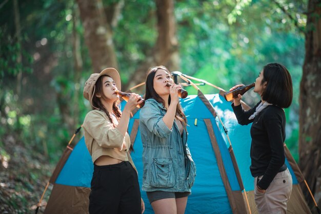 Gruppe schöner asiatischer Freundinnen, die sich vor dem Campingzelt entspannen. Sie genießen es, mit Spaß und Freude zusammen zu reden und Bier zu trinken