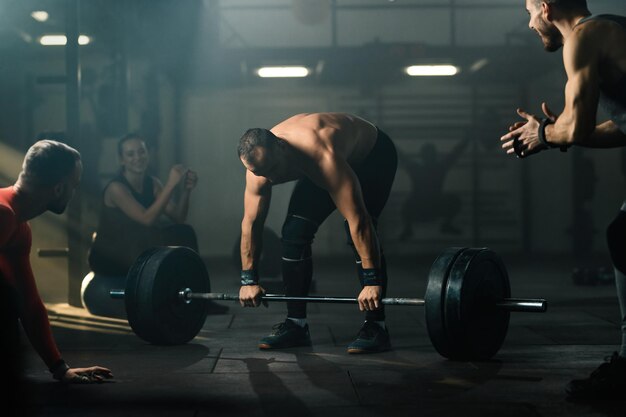 Gruppe von Athleten, die einen muskulösen Mann unterstützen, der Kreuzheben mit Langhantel in einem Fitnessstudio durchführt