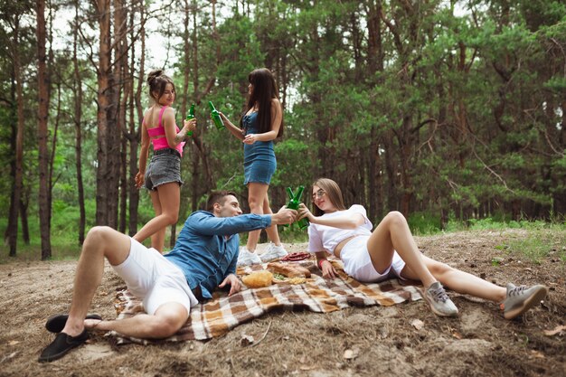 Gruppe von Freunden, die Bierflaschen während des Picknicks im Sommerwald klirren. Lebensstil, Freundschaft