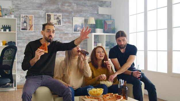 Gruppe von Freunden, die glücklich sind, nachdem ihre Lieblingsfußballmannschaft die Meisterschaft gewonnen hat. Freunde sitzen auf dem Sofa und essen Pizza und Pommes.