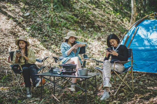 Gruppe von Freundinnen, die auf einem Campingstuhl sitzen und ihr Smartphone benutzen, während sie im Park campen und sich gegenseitig ignorieren