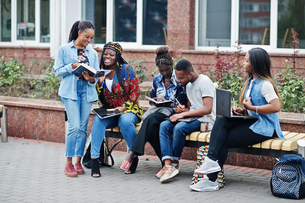 Gruppe von fünf afrikanischen College-Studenten, die gemeinsam Zeit auf dem Campus des Universitätshofs verbringen Schwarze Afro-Freunde, die an der Bank mit Schulartikeln, Laptops, Notizbüchern studieren