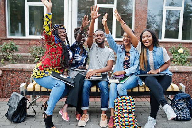 Gruppe von fünf afrikanischen College-Studenten, die gemeinsam Zeit auf dem Campus des Universitätshofs verbringen Schwarze Afro-Freunde, die an der Bank mit Schulartikeln, Laptops, Notizbüchern studieren