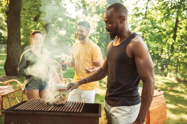 Gruppe von glücklichen Freunden, die Bier und Grillparty am sonnigen Tag haben. Zusammen im Freien in einer Waldlichtung oder im Hinterhof ausruhen