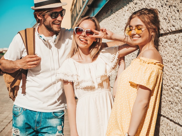 Gruppe von jungen drei stilvollen Freunden, die in der Straße aufwerfen. Modemann und zwei süße Mädchen gekleidet in lässige Sommerkleidung. Lächelnde Models, die Spaß an Sonnenbrillen haben. Fröhliche Frauen und Männer im Freien