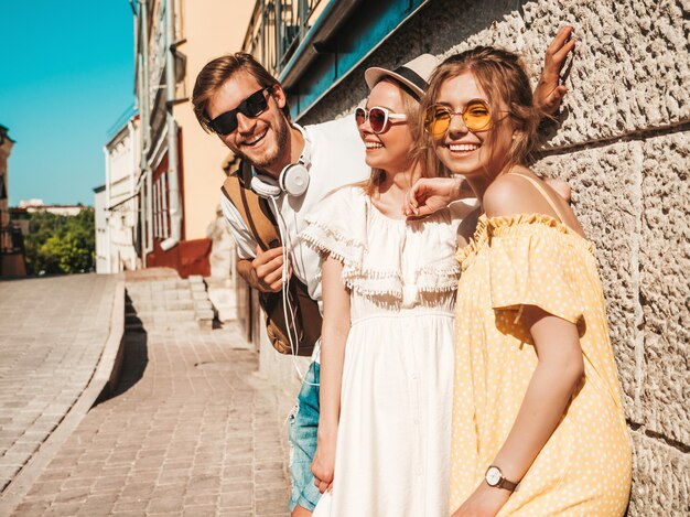 Gruppe von jungen drei stilvollen Freunden, die in der Straße aufwerfen. Modemann und zwei süße Mädchen gekleidet in lässige Sommerkleidung. Lächelnde Models, die Spaß an Sonnenbrillen haben. Fröhliche Frauen und Männer im Freien