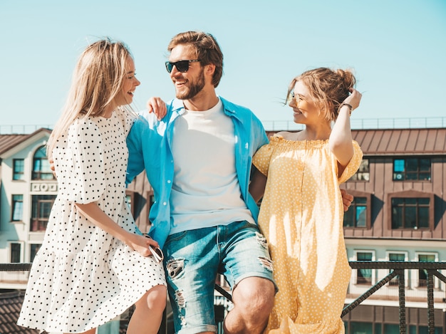 Gruppe von jungen drei stilvollen Freunden, die in der Straße aufwerfen. Modemann und zwei süße Mädchen gekleidet in lässige Sommerkleidung. Lächelnde Models, die Spaß an Sonnenbrillen haben. Fröhliche Frauen und Männer im Freien