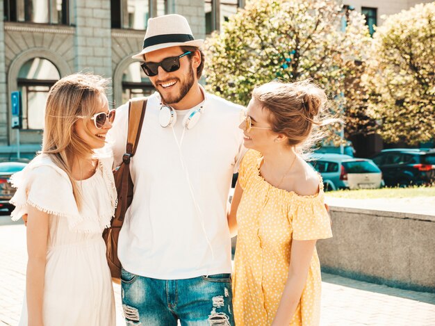 Gruppe von jungen drei stilvollen Freunden, die in der Straße aufwerfen. Modemann und zwei süße Mädchen gekleidet in lässige Sommerkleidung. Lächelnde Models, die Spaß an Sonnenbrillen haben. Fröhliche Frauen und Männer unterhalten sich