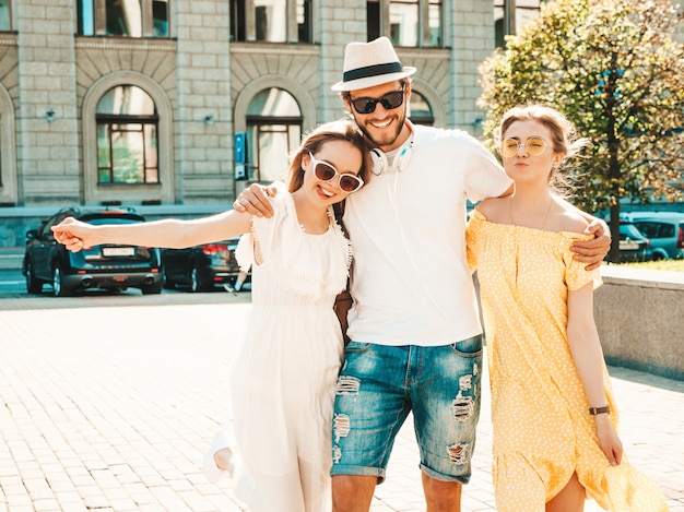 Gruppe von jungen drei stilvollen Freunden, die in der Straße aufwerfen. Modemann und zwei süße Mädchen gekleidet in lässige Sommerkleidung. Lächelnde Models, die Spaß an Sonnenbrillen haben. Vorsichtige Frauen und Kerle, die verrückt werden