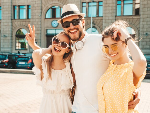 Gruppe von jungen drei stilvollen Freunden, die in der Straße aufwerfen. Modemann und zwei süße Mädchen gekleidet in lässige Sommerkleidung. Lächelnde Models, die Spaß an Sonnenbrillen haben. Vorsichtige Frauen und Kerle, die verrückt werden