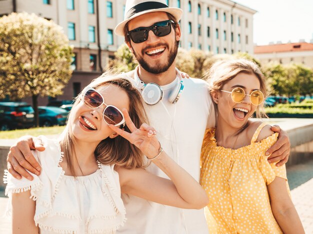 Gruppe von jungen drei stilvollen Freunden, die in der Straße aufwerfen. Modemann und zwei süße Mädchen gekleidet in lässige Sommerkleidung. Lächelnde Models, die Spaß an Sonnenbrillen haben. Vorsichtige Frauen und Kerle, die verrückt werden