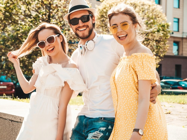 Gruppe von jungen drei stilvollen freunden, die in der straße aufwerfen. modemann und zwei süße mädchen gekleidet in lässige sommerkleidung. lächelnde models, die spaß an sonnenbrillen haben. vorsichtige frauen und kerle, die verrückt werden