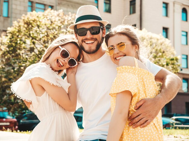 Gruppe von jungen drei stilvollen Freunden, die in der Straße aufwerfen. Modemann und zwei süße Mädchen gekleidet in lässige Sommerkleidung. Lächelnde Models, die Spaß an Sonnenbrillen haben. Vorsichtige Frauen und Kerle, die verrückt werden