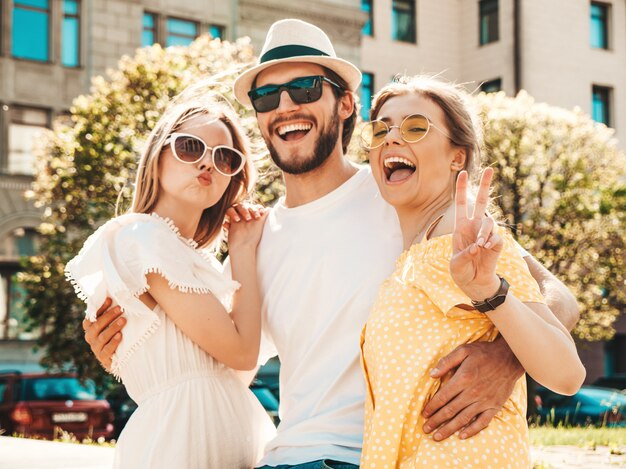 Gruppe von jungen drei stilvollen Freunden, die in der Straße aufwerfen. Modemann und zwei süße Mädchen gekleidet in lässige Sommerkleidung. Lächelnde Models, die Spaß an Sonnenbrillen haben. Vorsichtige Frauen und Kerle, die verrückt werden