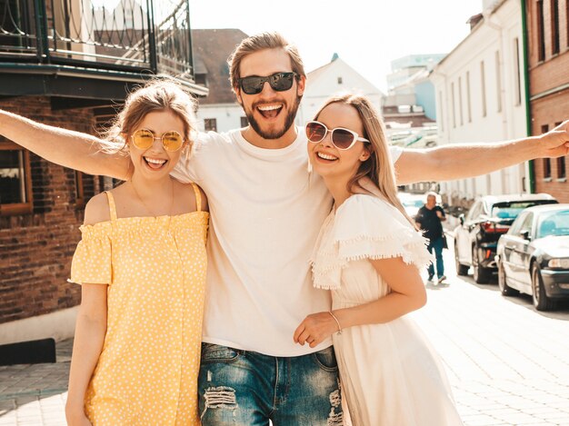 Gruppe von jungen drei stilvollen Freunden, die in der Straße aufwerfen. Modemann und zwei süße Mädchen gekleidet in lässige Sommerkleidung. Lächelnde Models, die Spaß an Sonnenbrillen haben. Vorsichtige Frauen und Kerle, die verrückt werden