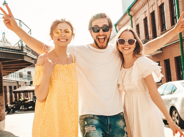 Gruppe von jungen drei stilvollen Freunden, die in der Straße aufwerfen. Modemann und zwei süße Mädchen gekleidet in lässige Sommerkleidung. Lächelnde Models, die Spaß an Sonnenbrillen haben. Vorsichtige Frauen und Kerle, die verrückt werden