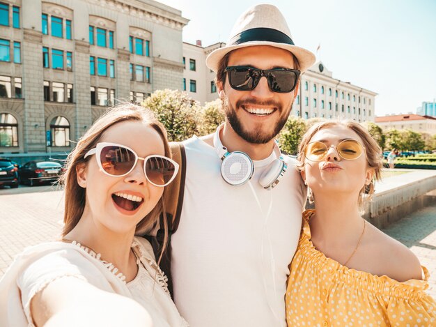 Gruppe von jungen drei stilvollen Freunden in der Straße. Mann und zwei süße Mädchen gekleidet in lässiger Sommerkleidung. Lächelnde Modelle, die Spaß in der Sonnenbrille haben. Frauen und Kerl, die Foto Selfie auf Smartphone machen