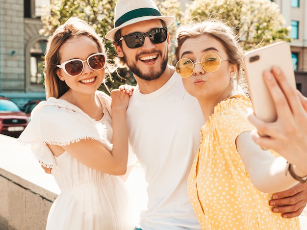 Gruppe von jungen drei stilvollen Freunden in der Straße. Mann und zwei süße Mädchen gekleidet in lässiger Sommerkleidung. Lächelnde Modelle, die Spaß in der Sonnenbrille haben. Frauen und Kerl, die Foto Selfie auf Smartphone machen