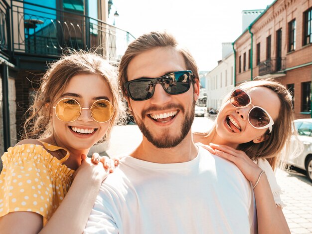 Gruppe von jungen drei stilvollen Freunden in der Straße. Mann und zwei süße Mädchen gekleidet in lässiger Sommerkleidung. Lächelnde Modelle, die Spaß in der Sonnenbrille haben. Frauen und Kerl, die Foto Selfie auf Smartphone machen