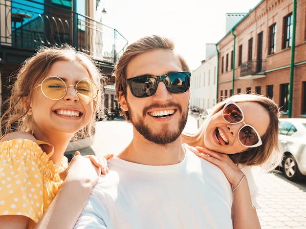 Gruppe von jungen drei stilvollen Freunden in der Straße. Mann und zwei süße Mädchen gekleidet in lässiger Sommerkleidung. Lächelnde Modelle, die Spaß in der Sonnenbrille haben. Frauen und Kerl, die Foto Selfie auf Smartphone machen