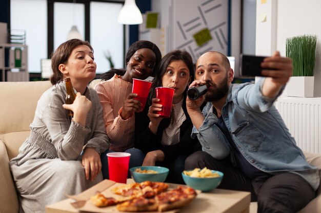 Gruppe von Menschen, die Selfies machen und nach Feierabend mit Getränken im Büro feiern. Mann und Frau haben Spaß mit Bildern auf dem Smartphone und genießen alkoholische Getränke und Snacks auf der Party.