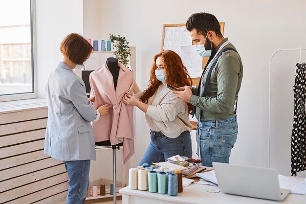 Gruppe von Modedesignern mit medizinischen Masken, die im Atelier arbeiten und Kleidungsstück auf Kleidform überprüfen