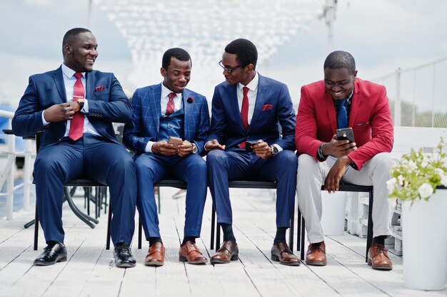 Gruppe von vier afroamerikanischen glücklichen erfolgreichen Männern im Anzug Reiche schwarze Geschäftsleute sitzen auf Stühlen und schauen auf Mobiltelefone